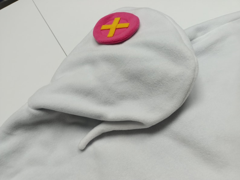 May include: White fleece hat with a pink cross emblem. The hat has a rounded shape with a pointed tail. The cross emblem is a pink circle with a yellow cross in the center. The hat is made of a soft, plush material.