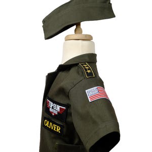 May include: Olive green Top Gun Maverick costume for children, including a jacket, hat, and name tag. The jacket features the American flag patch and the name "Oliver". The costume is designed to resemble a military uniform.