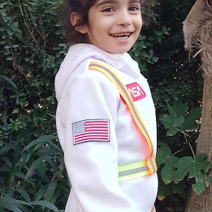 Op de afbeelding: Een jong kind draagt een wit astronautenkostuum met een rood, wit en blauwe Amerikaanse vlagpatch op de mouw en een gele en oranje reflecterende band over de borst. Het kostuum heeft het woord "NASA" op de borst gedrukt. Het kind glimlacht en kijkt naar de camera.