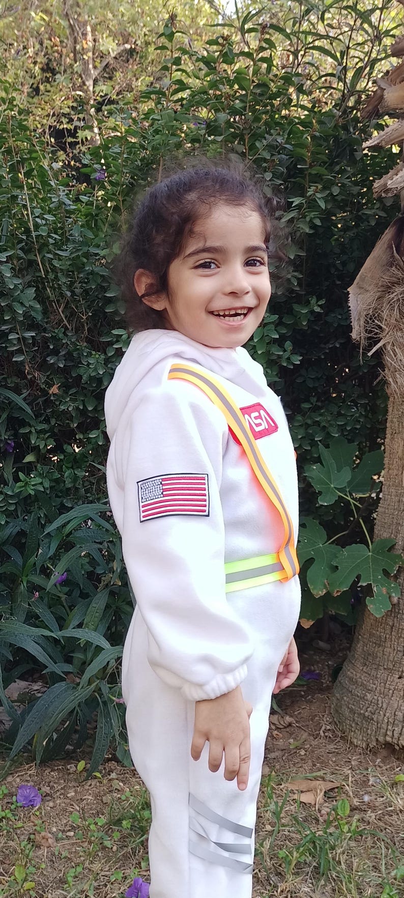 Op de afbeelding: Een jong kind draagt een wit astronautenkostuum met een rood, wit en blauwe Amerikaanse vlagpatch op de mouw en een gele en oranje reflecterende band over de borst. Het kostuum heeft het woord "NASA" op de borst gedrukt. Het kind glimlacht en kijkt naar de camera.