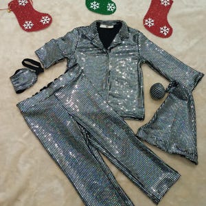 May include: A silver sequined pajama set with a collared shirt, pants, a face mask, and a sleep eye mask. The set is displayed with red and green Christmas stockings with white snowflake designs.