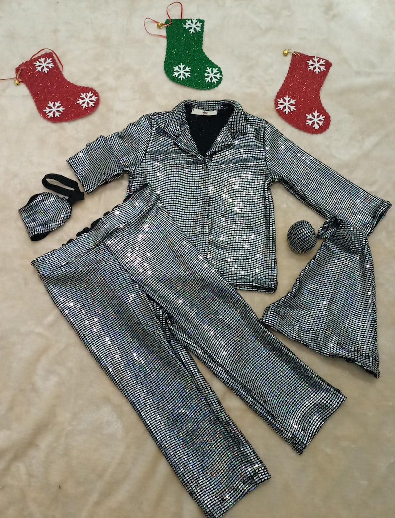 May include: A silver sequined pajama set with a collared shirt, pants, a face mask, and a sleep eye mask. The set is displayed with red and green Christmas stockings with white snowflake designs.
