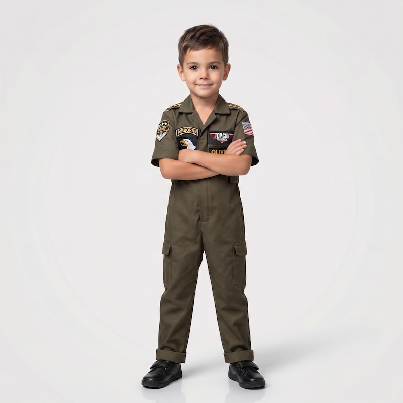 May include: A child wearing an olive green Top Gun flight suit costume. The short-sleeved jumpsuit features patches, including an American flag and the word "Airborne." The child is wearing black shoes and has their arms crossed.