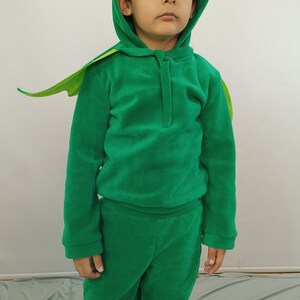 May include: A child wearing a green fleece dinosaur costume with a hood and a tail. The costume has a zipper in the front and a green hood with a pointed top.