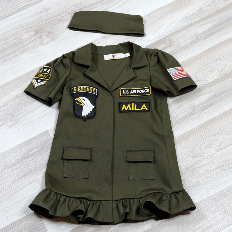 Puede incluir: Un vestido verde de estilo militar con parches que dicen "Airborne", "U.S. Air Force" y "Mila". El vestido tiene un dobladillo con volantes y viene con una diadema verde a juego.
