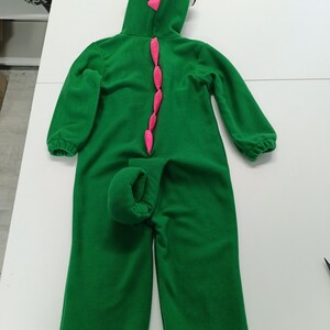 May include: Green dinosaur costume with a hood, pink accents, and a tail. The hooded jumpsuit features a row of pink spikes down the back and a curled tail. The hood has yellow and pink details. The costume is made of a soft material.