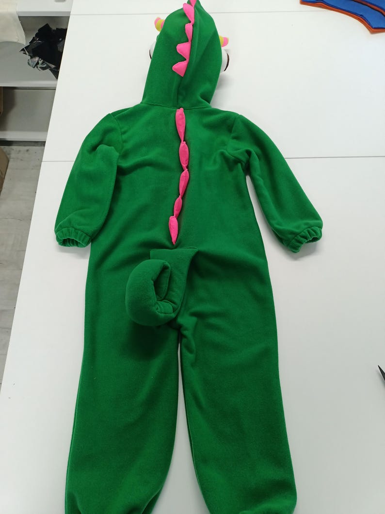 May include: Green dinosaur costume with a hood, pink accents, and a tail. The hooded jumpsuit features a row of pink spikes down the back and a curled tail. The hood has yellow and pink details. The costume is made of a soft material.