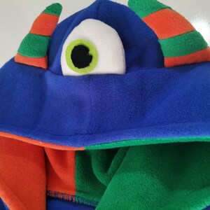 May include: A blue monster hood with an orange and green striped horn and a large white eye with a green iris and black pupil. The hood's interior is orange and green.