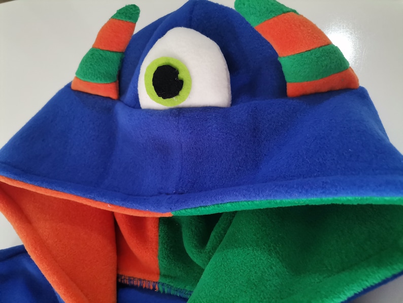 May include: A blue monster hood with an orange and green striped horn and a large white eye with a green iris and black pupil. The hood's interior is orange and green.