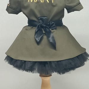 May include: Olive green dress with a black tulle skirt and a black satin bow at the waist. The name "MARY" is embroidered in gold on the back of the dress. The dress is displayed on a wooden stand.