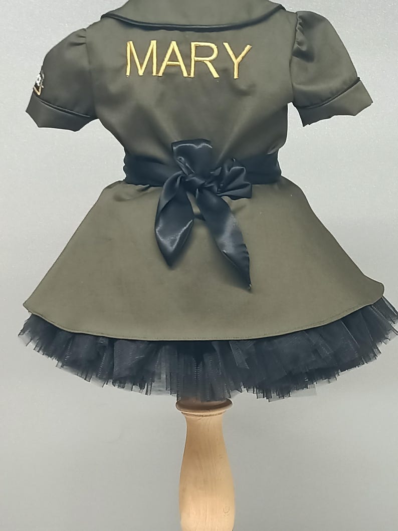 May include: Olive green dress with a black tulle skirt and a black satin bow at the waist. The name "MARY" is embroidered in gold on the back of the dress. The dress is displayed on a wooden stand.