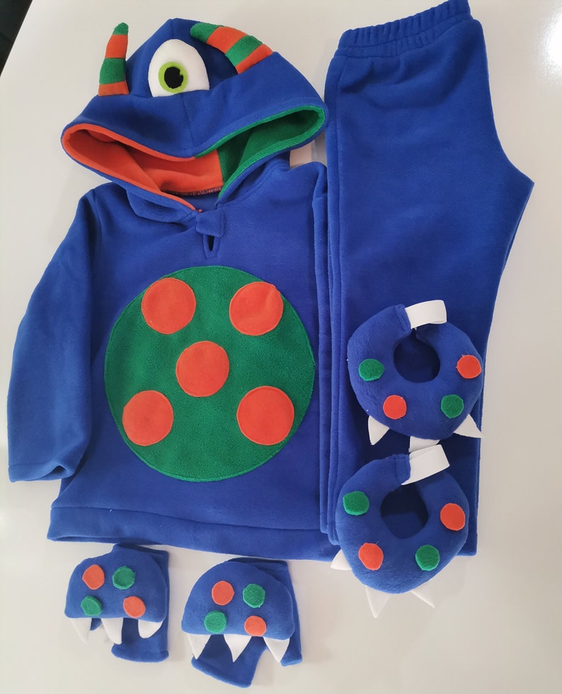 May include: A blue monster-themed costume set for children. The set includes a hooded sweatshirt with a large green and orange circle design, matching pants, and monster feet and hand covers. The hood has a large eye and horns.