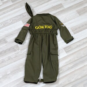 May include: Olive green jumpsuit with the word "G&Ouml;KTUĞ" in yellow lettering on the back. The costume features a US flag patch on the left sleeve and a matching hat. The jumpsuit has multiple pockets and a military-style patch.