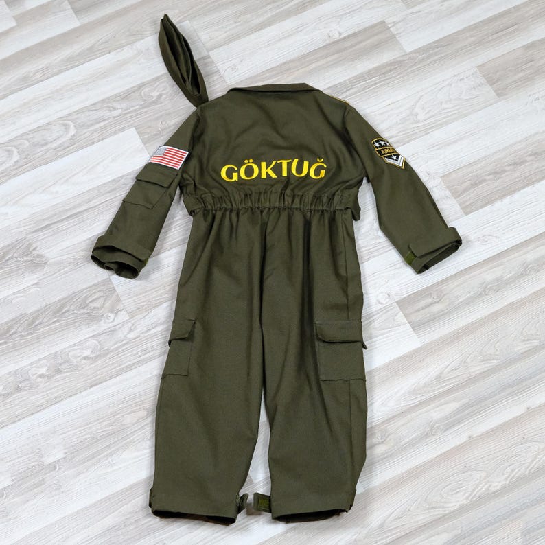 May include: Olive green jumpsuit with the word "G&Ouml;KTUĞ" in yellow lettering on the back. The costume features a US flag patch on the left sleeve and a matching hat. The jumpsuit has multiple pockets and a military-style patch.