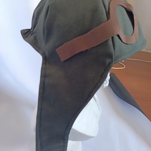 May include: A dark olive green aviator hat with brown felt goggles attached. The hat has ear flaps and is displayed on a white bust. The goggles are a key feature, adding to the vintage aesthetic. The hat is made of a soft fabric.