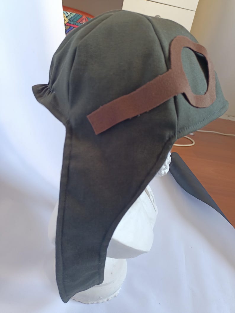 May include: A dark olive green aviator hat with brown felt goggles attached. The hat has ear flaps and is displayed on a white bust. The goggles are a key feature, adding to the vintage aesthetic. The hat is made of a soft fabric.