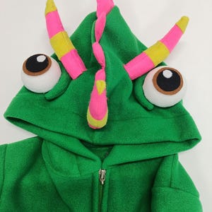 May include: A green hooded costume with large, cartoonish eyes and horn-like protrusions. The horns are pink and yellow, and the eyes are white with brown and black details. The costume has a zipper closure.