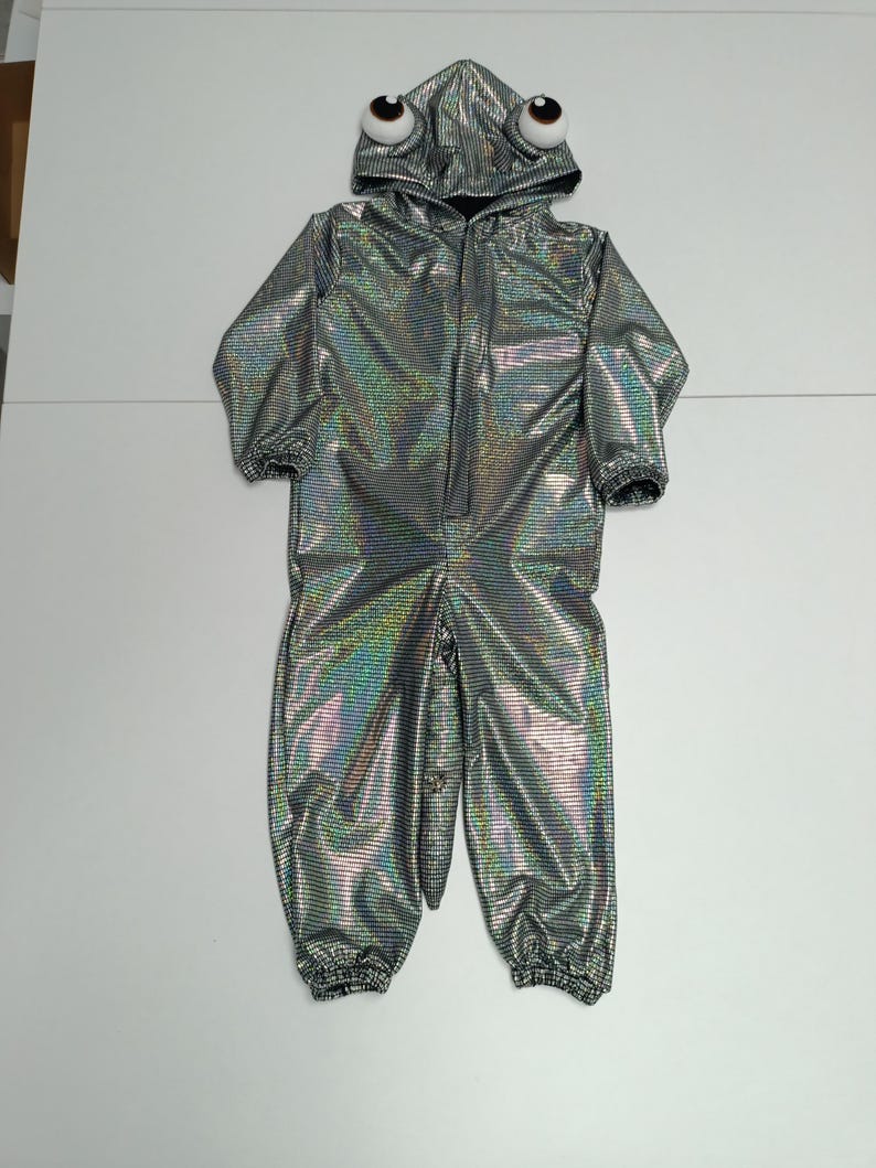 K&ouml;nnte beinhalten: Ein silberner, schillernder Alien-Anzug mit Kapuze und gro&szlig;en, cartoonartigen Augen. Der einteilige Anzug hat einen Rei&szlig;verschluss und gerippte B&uuml;ndchen an Handgelenken und Kn&ouml;cheln. Das Kost&uuml;m ist f&uuml;r Kinder und aus gl&auml;nzendem Stoff.