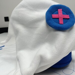 May include: A white fleece hat with a blue circular emblem featuring a pink cross. The hat has a pointed shape and is likely a costume piece. The hat is resting on a blue cylindrical object.
