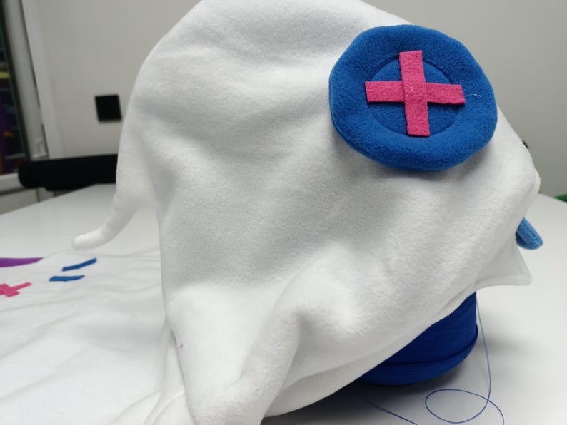 May include: A white fleece hat with a blue circular emblem featuring a pink cross. The hat has a pointed shape and is likely a costume piece. The hat is resting on a blue cylindrical object.