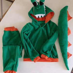 Peut inclure: Costume de dinosaure vert et orange avec un sweat &agrave; capuche, un pantalon et une queue. La t&ecirc;te de dinosaure est attach&eacute;e &agrave; la capuche.