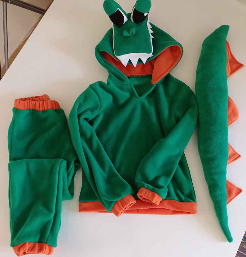 Peut inclure: Costume de dinosaure vert et orange avec un sweat &agrave; capuche, un pantalon et une queue. La t&ecirc;te de dinosaure est attach&eacute;e &agrave; la capuche.