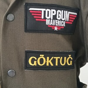 Puede incluir: Chaqueta verde oliva de Top Gun Maverick con el texto "TOP GUN MAVERICK" y una estrella en un parche negro. Debajo, un parche con el nombre "G&Ouml;KTUĞ" en letras amarillas.