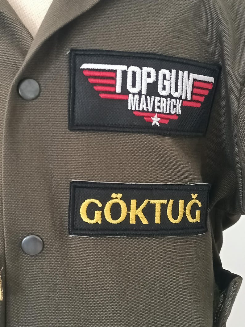 Puede incluir: Chaqueta verde oliva de Top Gun Maverick con el texto "TOP GUN MAVERICK" y una estrella en un parche negro. Debajo, un parche con el nombre "G&Ouml;KTUĞ" en letras amarillas.