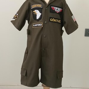 Puede incluir: Mono verde oliva con m&uacute;ltiples parches, incluyendo "AIRBORNE" con un &aacute;guila, "TOP GUN MAVERICK" y "U.S. AIR FORCE". El mono de manga corta tiene bolsillos y el nombre "G&Ouml;KTUĞ" en el pecho.