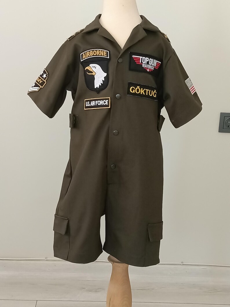 Puede incluir: Mono verde oliva con m&uacute;ltiples parches, incluyendo "AIRBORNE" con un &aacute;guila, "TOP GUN MAVERICK" y "U.S. AIR FORCE". El mono de manga corta tiene bolsillos y el nombre "G&Ouml;KTUĞ" en el pecho.
