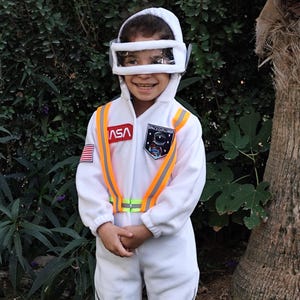 Op de afbeelding: Een kind in een wit astronautenkostuum met een oranje en gele reflecterende vest. Het kostuum heeft een NASA-patch en de tekst "Space Explorer".
