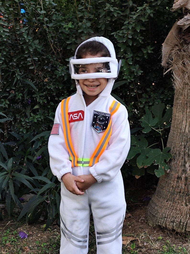 Op de afbeelding: Een kind in een wit astronautenkostuum met een oranje en gele reflecterende vest. Het kostuum heeft een NASA-patch en de tekst "Space Explorer".