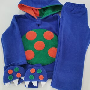 May include: A blue monster-themed fleece outfit, including a hooded sweatshirt, pants, and monster claw mittens. The sweatshirt features a green and orange design with orange spots and a large eye on the hood.