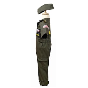 May include: Olive green pilot costume for children, including a jumpsuit and cap. The jumpsuit features patches, an American flag, and the name "OLIVER". The cap sits atop a wooden mannequin head. The costume has pockets and yellow lettering.