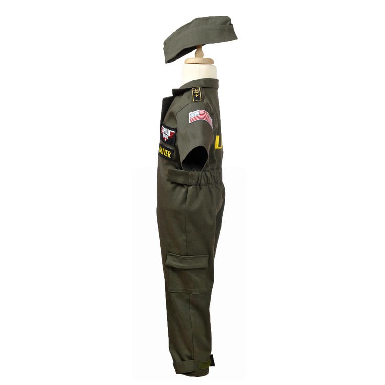 May include: Olive green pilot costume for children, including a jumpsuit and cap. The jumpsuit features patches, an American flag, and the name "OLIVER". The cap sits atop a wooden mannequin head. The costume has pockets and yellow lettering.