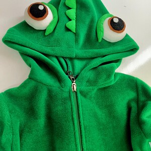 May include: Green fleece hooded jacket with a zipper closure and a chameleon design on the hood. The hood has large, white eyes with black pupils and a green, scaly, chameleon head.