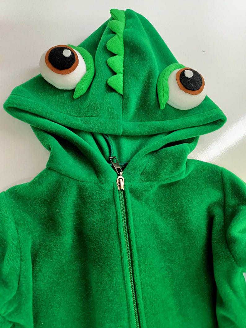 May include: Green fleece hooded jacket with a zipper closure and a chameleon design on the hood. The hood has large, white eyes with black pupils and a green, scaly, chameleon head.