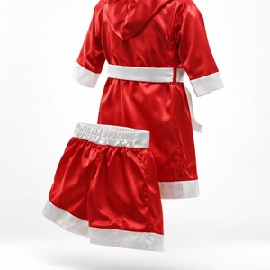 May include: Red satin boxing robe and shorts set with white trim. The robe has a hood, a white belt, and white cuffs. The shorts have a white waistband and white trim on the legs. The set is designed for a child.