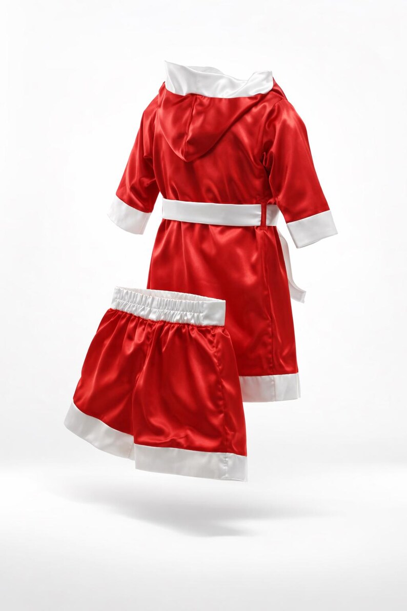 May include: Red satin boxing robe and shorts set with white trim. The robe has a hood, a white belt, and white cuffs. The shorts have a white waistband and white trim on the legs. The set is designed for a child.