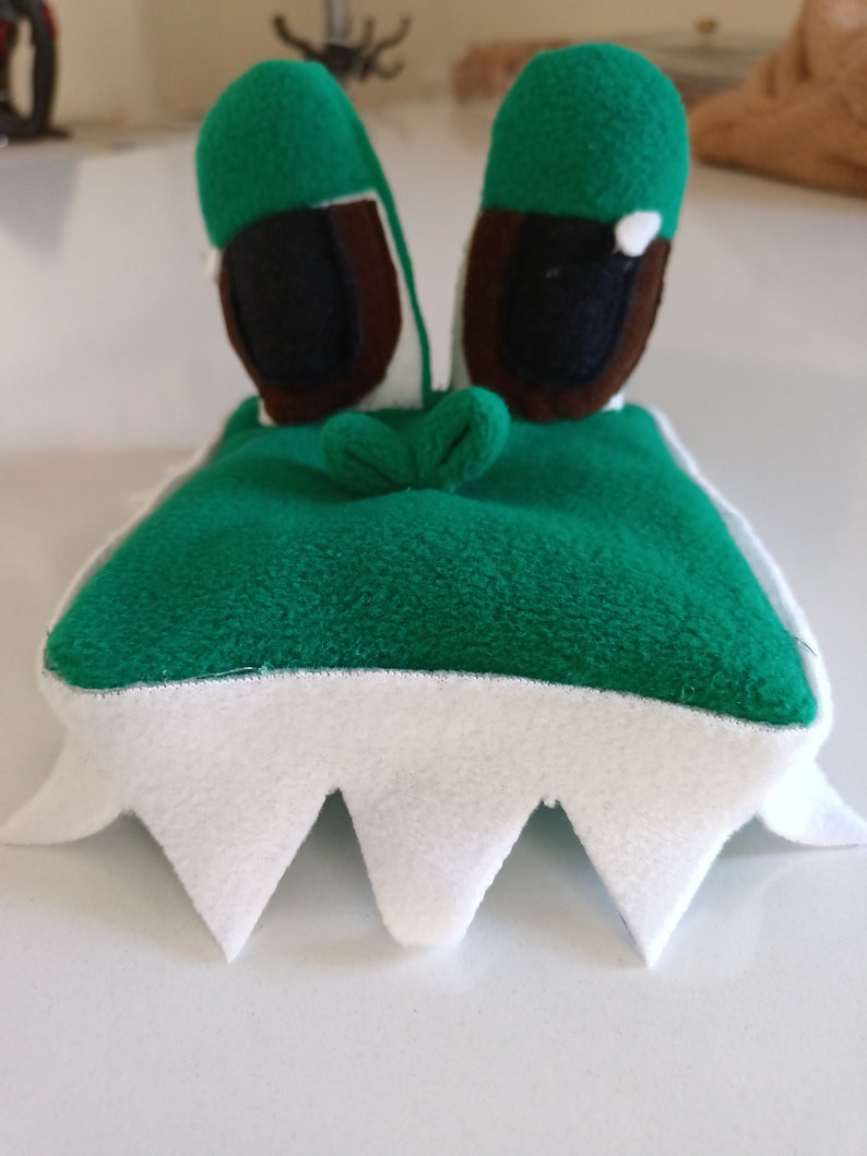 Peut inclure: Jouet en peluche vert avec des dents blanches et des yeux bruns. Le jouet a la forme d'un monstre avec une grosse t&ecirc;te et un petit corps.