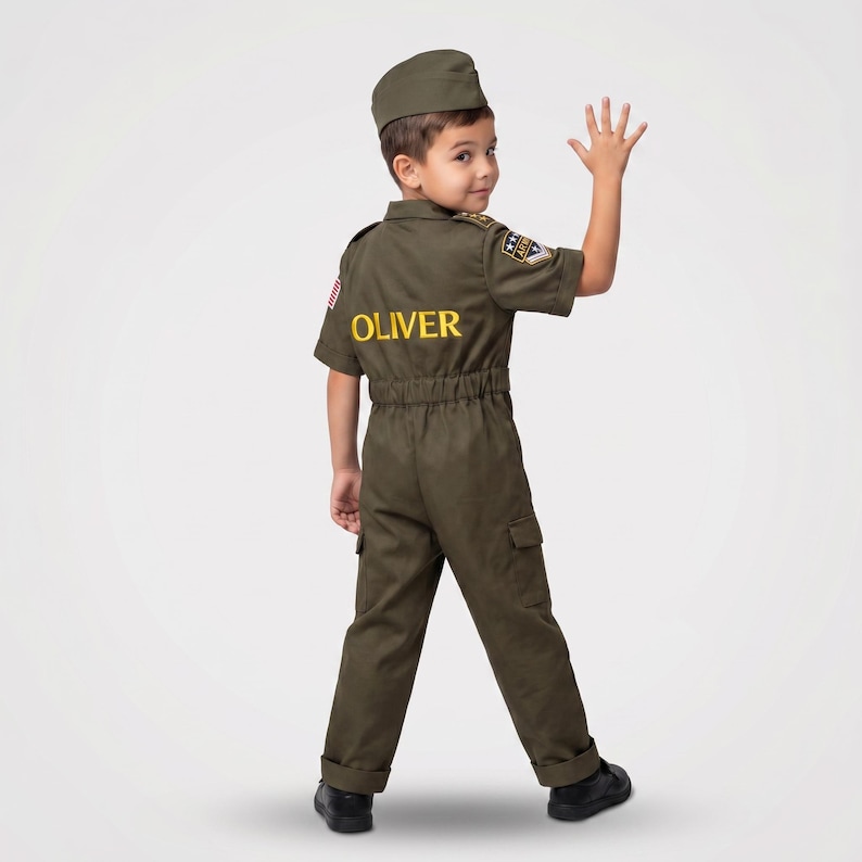 May include: A child wearing an olive green pilot costume, including a cap and jumpsuit. The jumpsuit has the name "OLIVER" in yellow lettering on the back. The child is waving with their right hand.