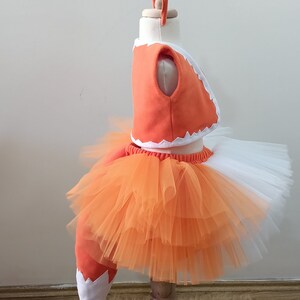 May include: Orange and white fox costume for a child. The costume includes a tulle skirt, a top with a white trim, and a tail.