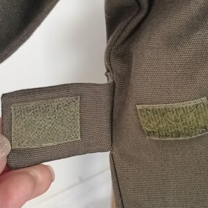 Puede incluir: Primer plano de una secci&oacute;n de una bolsa de lona verde oliva. La bolsa presenta un cierre de velcro rectangular en una correa, con un parche de velcro a juego en el cuerpo principal. La tela tiene un tejido texturizado.