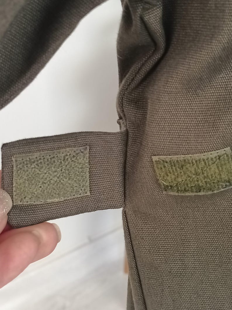 Puede incluir: Primer plano de una secci&oacute;n de una bolsa de lona verde oliva. La bolsa presenta un cierre de velcro rectangular en una correa, con un parche de velcro a juego en el cuerpo principal. La tela tiene un tejido texturizado.