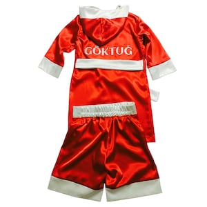 May include: A red satin boxing robe and shorts set with white trim. The robe has a hood and the name "G&Ouml;KTUĞ" embroidered in white. The shorts have a white waistband and trim. The set is designed for boxing.