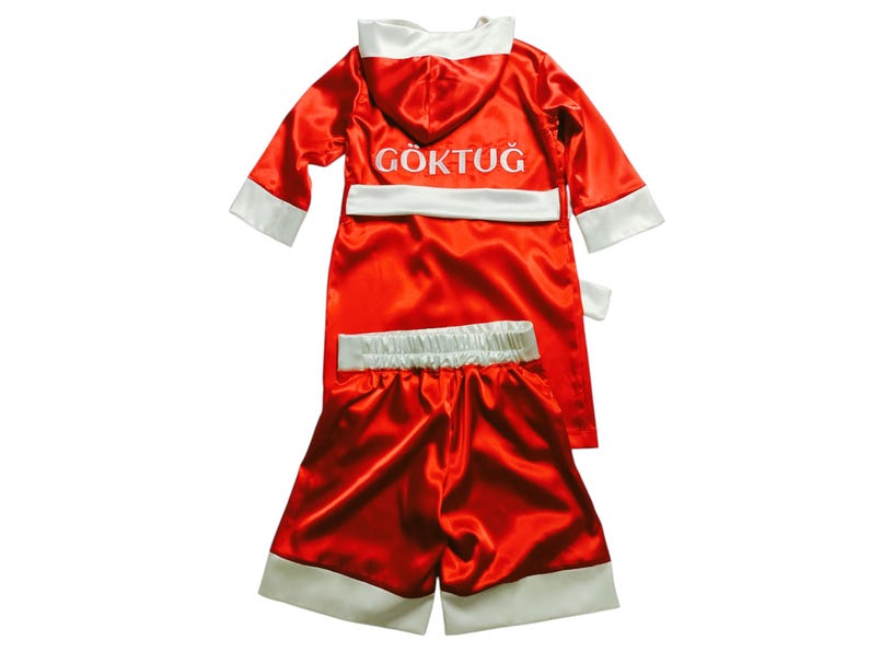 May include: A red satin boxing robe and shorts set with white trim. The robe has a hood and the name "G&Ouml;KTUĞ" embroidered in white. The shorts have a white waistband and trim. The set is designed for boxing.