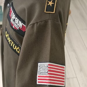 Puede incluir: Chaqueta verde oliva con parches que incluyen los textos "TOP GUN MAVERICK" y "G&Ouml;KTUĞ", y un parche de la bandera estadounidense. La chaqueta tiene cuello e insignias de estrellas doradas en el hombro.