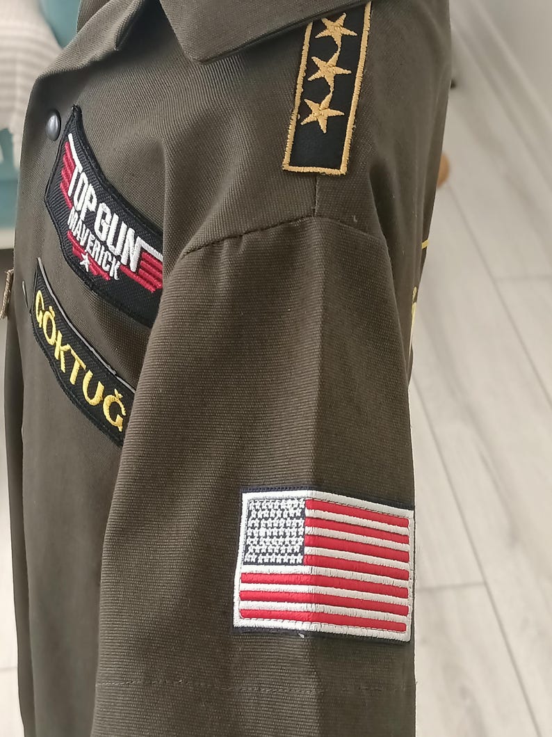 Puede incluir: Chaqueta verde oliva con parches que incluyen los textos "TOP GUN MAVERICK" y "G&Ouml;KTUĞ", y un parche de la bandera estadounidense. La chaqueta tiene cuello e insignias de estrellas doradas en el hombro.
