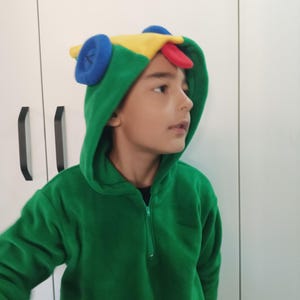 Puede incluir: Un ni&ntilde;o lleva una sudadera con capucha de color verde de forro polar con una capucha de personaje colorido. La capucha tiene un dise&ntilde;o amarillo, rojo, azul y verde.