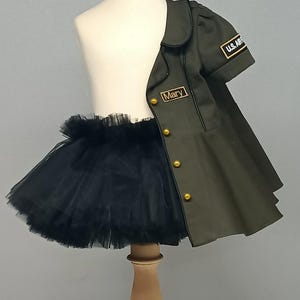 May include: A child's olive green jacket with gold buttons and "U.S. ARMY" patch, paired with a black tulle tutu. The jacket has the name "Mary" embroidered on it. The outfit is displayed on a mannequin.